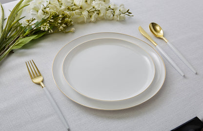 A place setting on a white tablecloth featuring a white plastic dinner plate with a delicate gold rim, accompanied by a gold-finished fork, knife, and spoon with white handles, styled with white flowers.