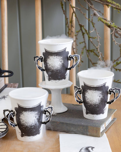 Paper cups with black cauldron design with paper handles on a wooden surface with books and branches in the background.