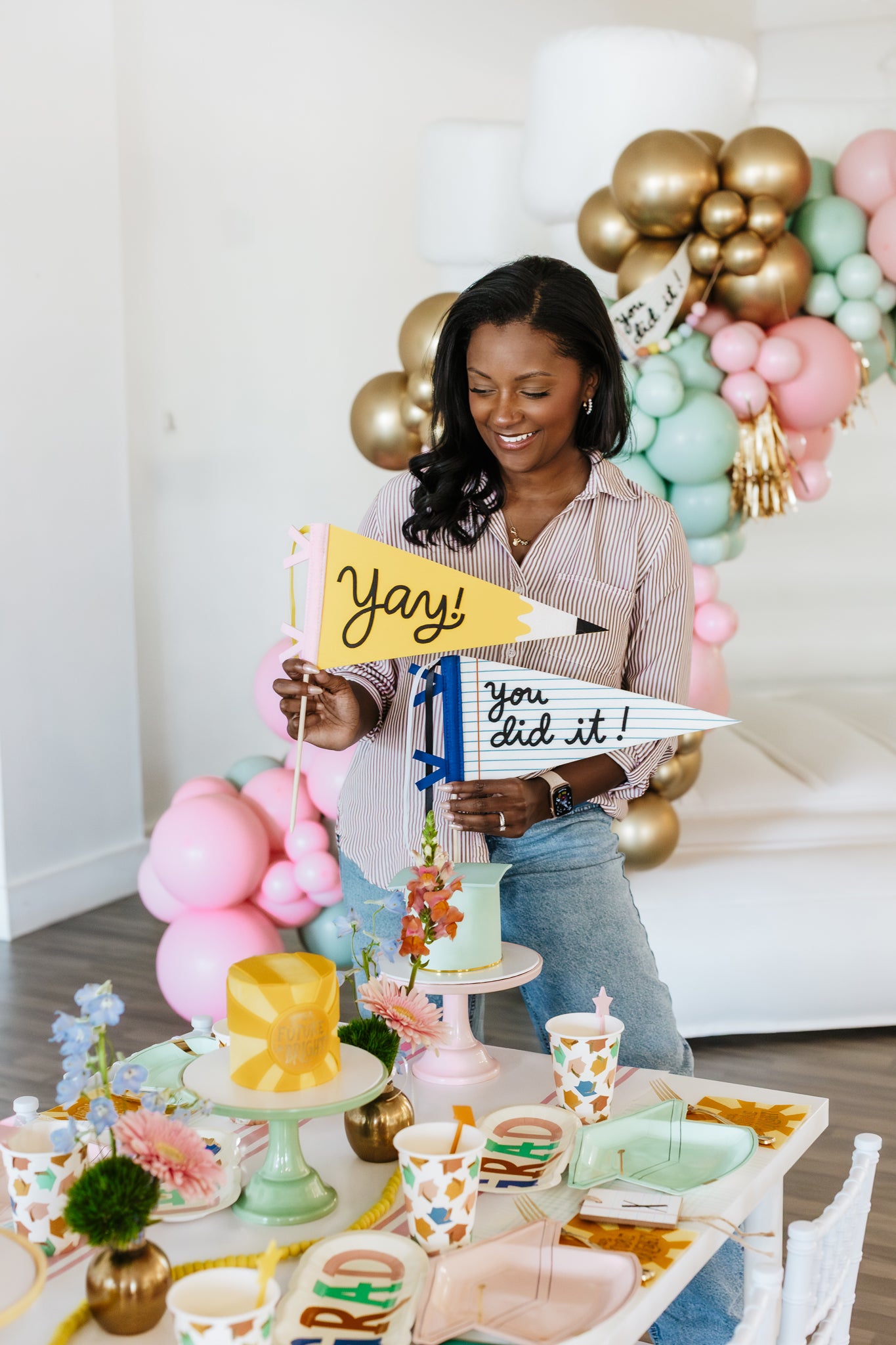 Woman holding 'yay! you did it!' graduation sign in a decorated room with balloons and cake.