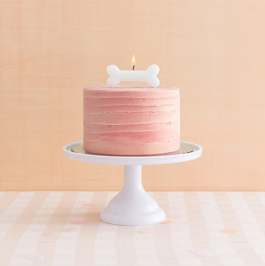 A large bone-shaped candle on top of a pink frosted cake, placed on a white stand against a beige background.