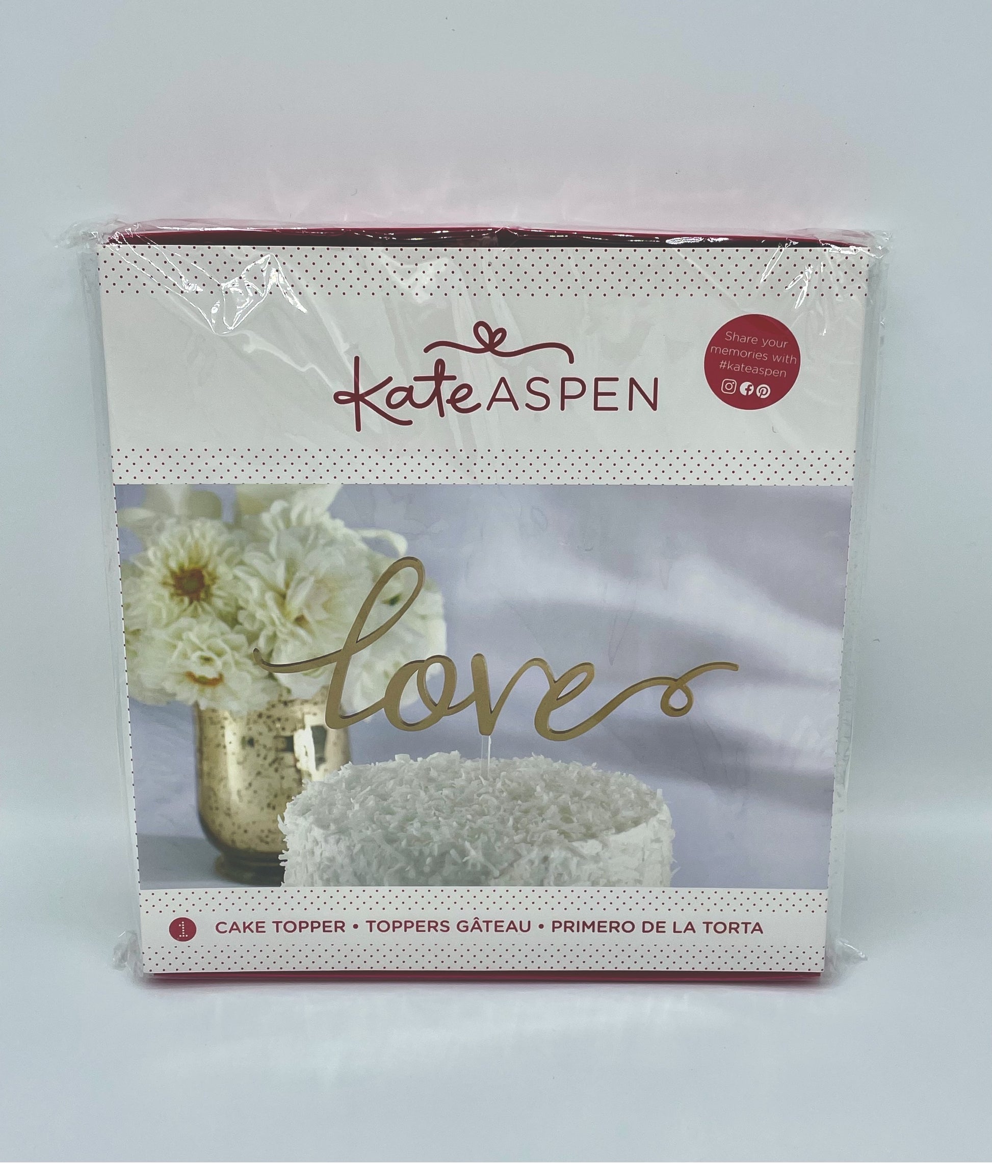 Kate Aspen 'Love' cake topper in its original retail packaging, featuring a gold script topper on a white cake, with a photo of white flowers in a gold vase.