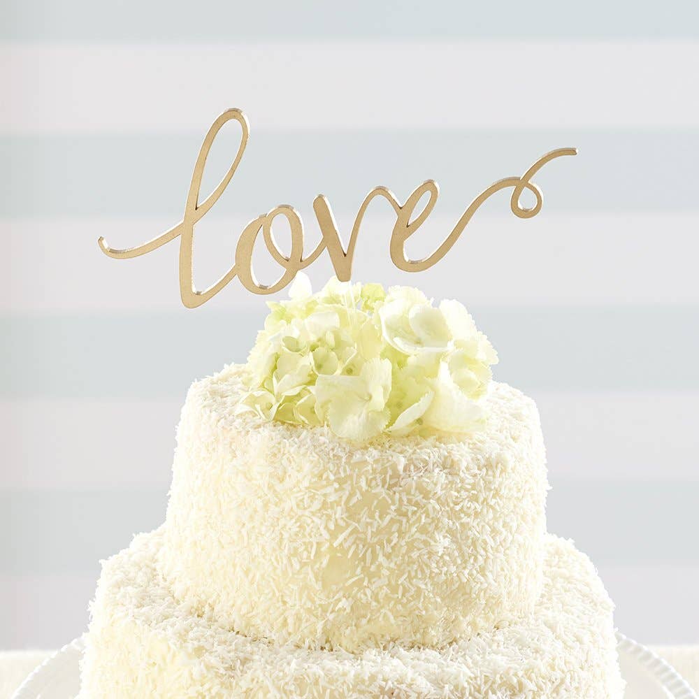 A gold acrylic cake topper spelling out the word 'love' in cursive script, placed on a white cake.