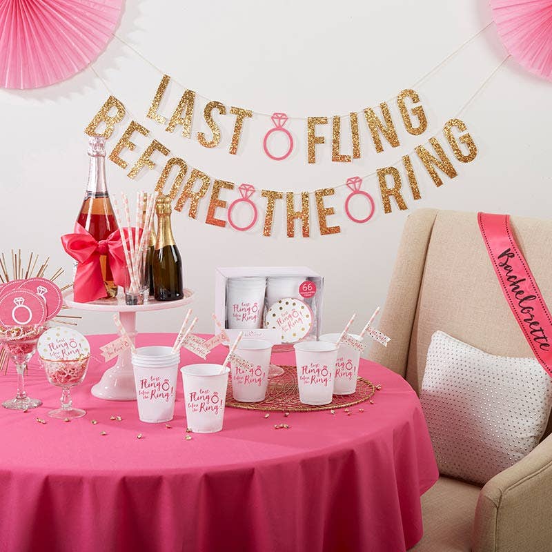 Bachelorette party supplies including pink and gold themed decorations, cups, and paper coasters displayed on a table with a pink tablecloth.