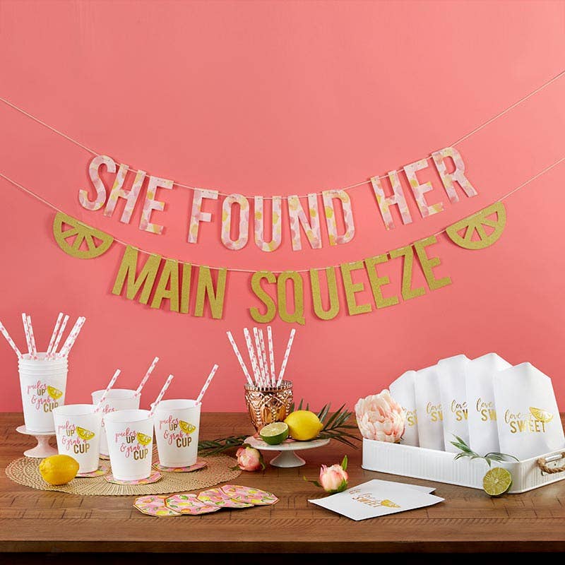 A party setup featuring a banner reading 'SHE FOUND HER MAIN SQUEEZE', white stadium cups with lemon slice design, gold foil trim hexagon coasters, paper straws, and cookie bags with lemon accents.