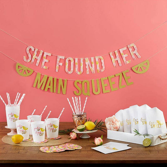 A party setup featuring a banner reading 'SHE FOUND HER MAIN SQUEEZE', white stadium cups with lemon slice design, gold foil trim hexagon coasters, paper straws, and cookie bags with lemon accents.