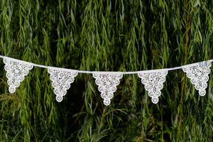 White Paper Lace Pennant Party Garland featuring white ribbon, set against a natural, green botanical leaf background