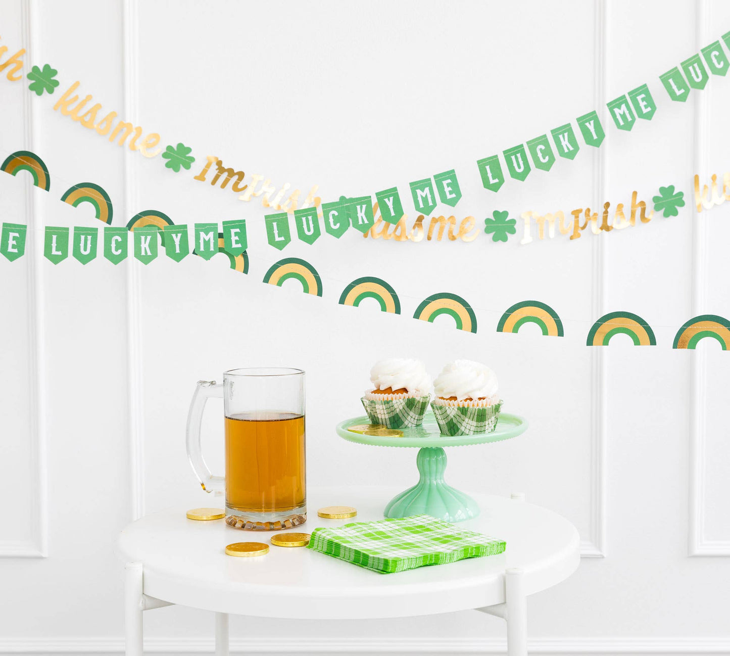 St. Patrick's Day themed banners with the text 'LUCKY ME' and 'KISS ME I'M IRISH', as well as green / gold rainbow banner, displayed on a table with a green cup and cupcakes.
