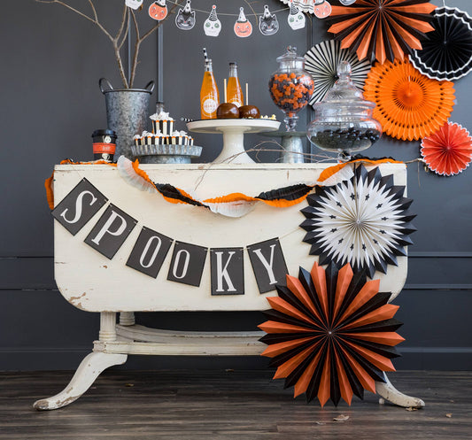 A 'SPOOKY' word banner with black glitter accents, designed for Halloween decorations, displayed over a table with themed items.