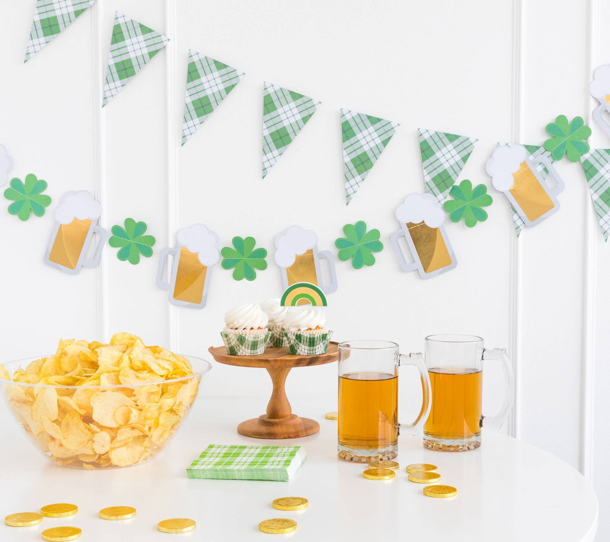 A festive St. Patrick's Day banner set with gold foiled mugs and shamrocks and one with green plaid pennants, displayed over a table with snacks and drinks.
