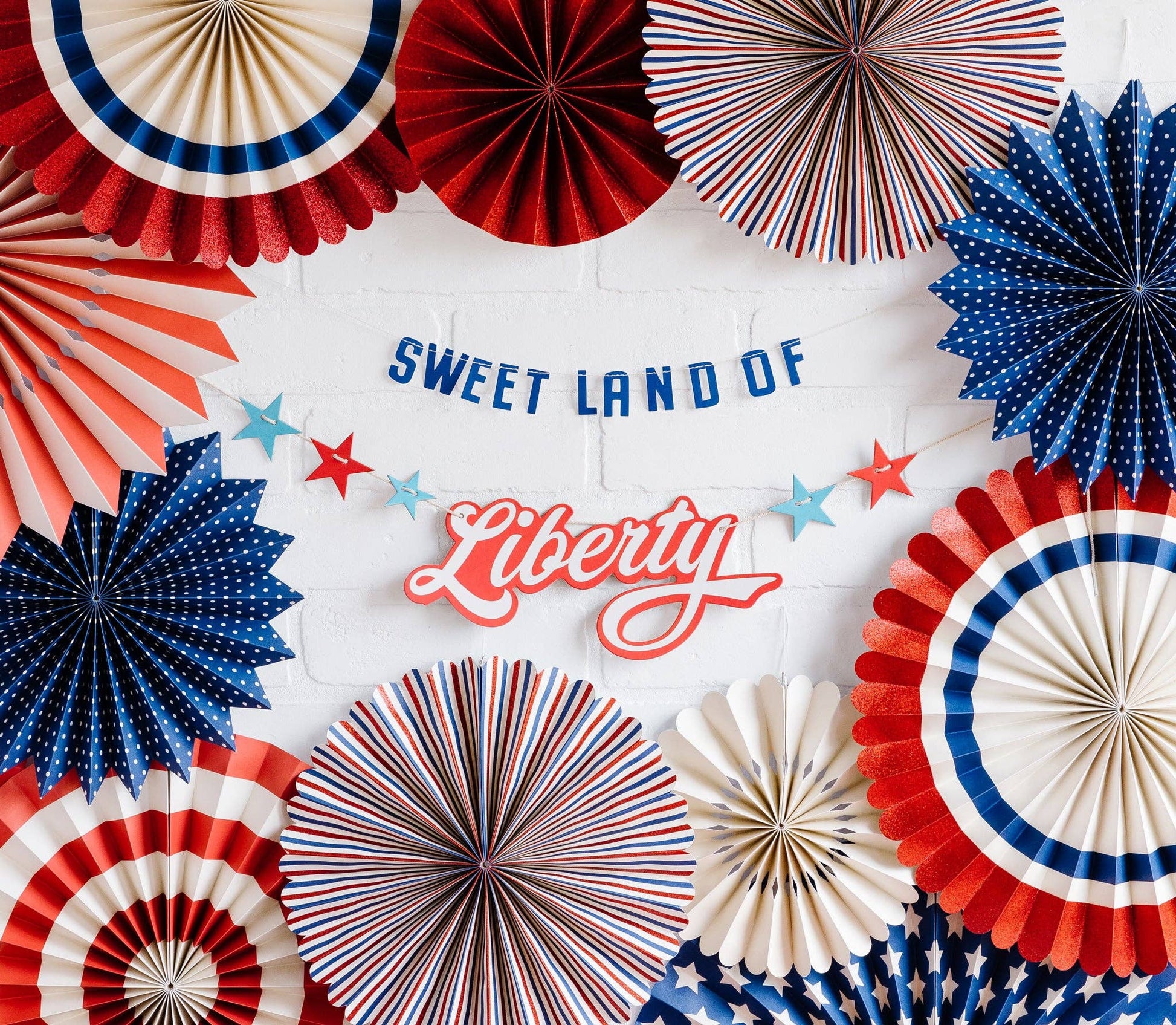 A set of paper fan decorations and banners with the text 'SWEET LAND OF Liberty' displayed prominently among them, decorated with red, white, and blue colors.
