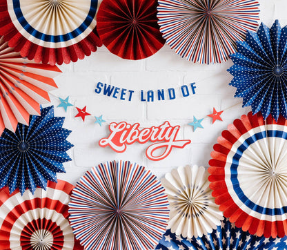 A set of paper fan decorations and banners with the text 'SWEET LAND OF Liberty' displayed prominently among them, decorated with red, white, and blue colors.